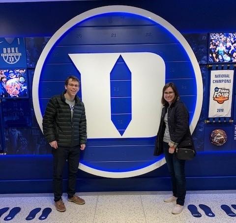 man and woman smiling in front of a blue wall with the Duke "D"