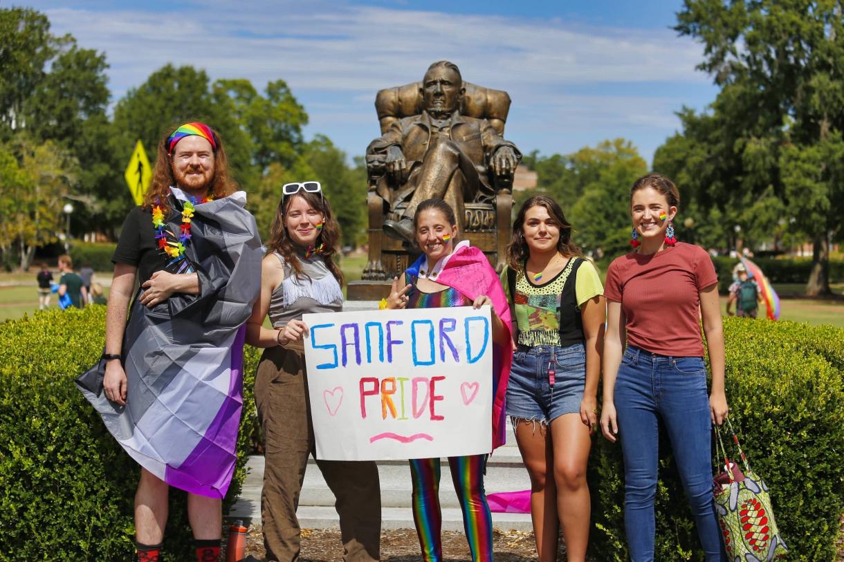 Students in rainbow colors pose with Sanford Pride flag in front of Duke statue