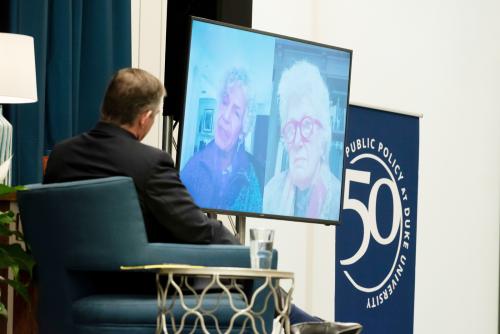 Two women on large screen talking to man in chair.