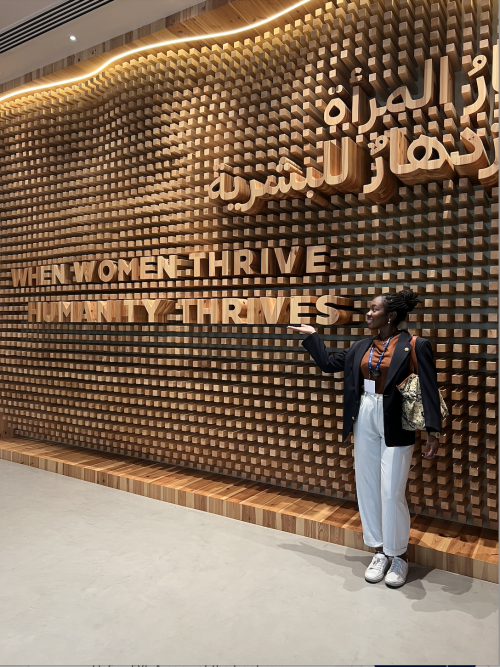 Eni Owoeye posing in front of an art installation that with the message "when women thrive, humanity thrives"