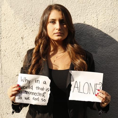 Woman standing in black clothing with two signs in her hands. One says " Why in a world that is so "connected do we feel so alone" and the other says "alone"/