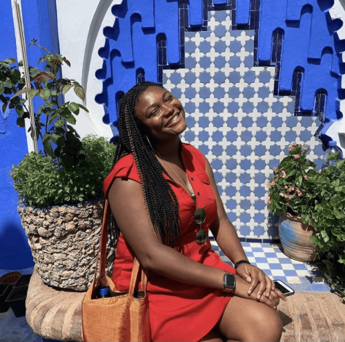 Woman smiling in red dress, sitting down in between plants with blue wall behind.