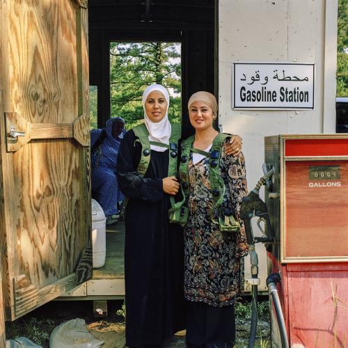 Two women, wearing headscarves, pose near building and gas station